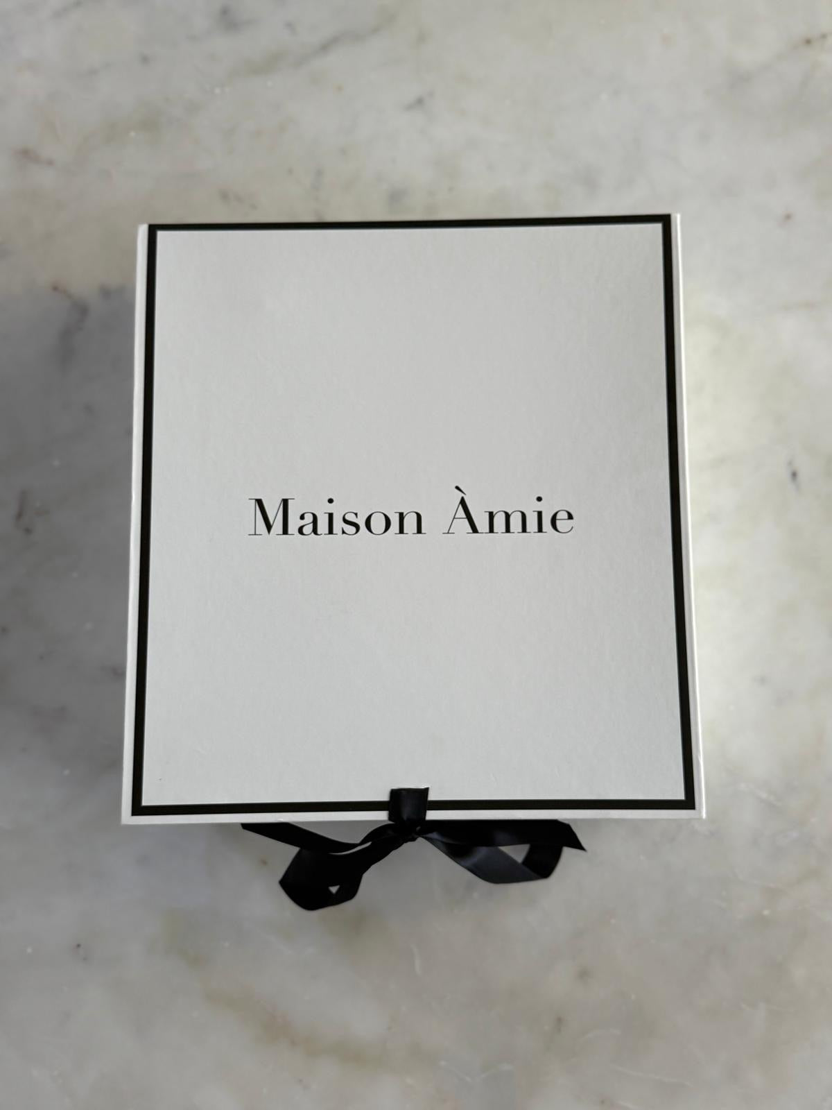 A white Maison Àmie gift box with a black border, black printed logo text, and a black ribbon bow at the bottom, placed on a light marble surface.