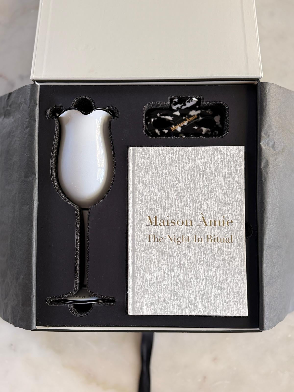 A boxed gift set from Maison Àmie titled 'The Night In Ritual,' shown open on a light marble surface. Inside the Maison Àmie Friendship Box - Fill-Me-In glass, Night-In Ritual journal and infinity hair clip nestled in the box and the bracelet under the journal.